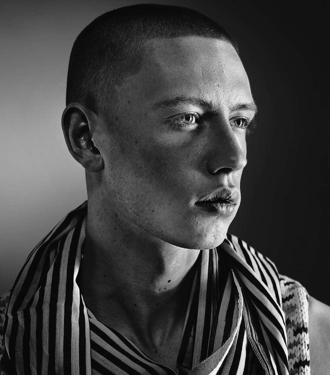 Portrait of a young adult male with short hair wearing a striped shirt. The image is black and white and the man is looking off to the side.