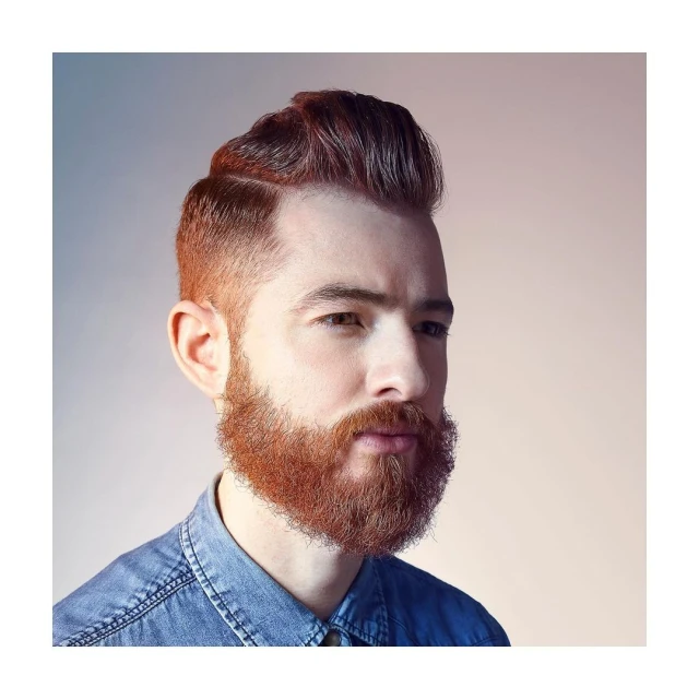 Portrait of a red-haired bearded man with a side part wearing a denim shirt against a neutral background in a head and shoulders shot.