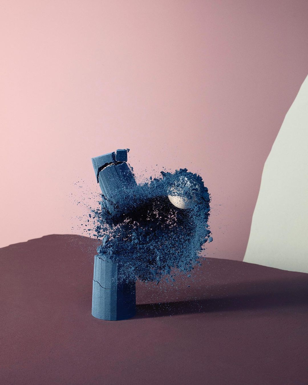 Broken cylinder with blue powder explosion around a sphere against a pink backdrop with geometric shapes and a shadow.