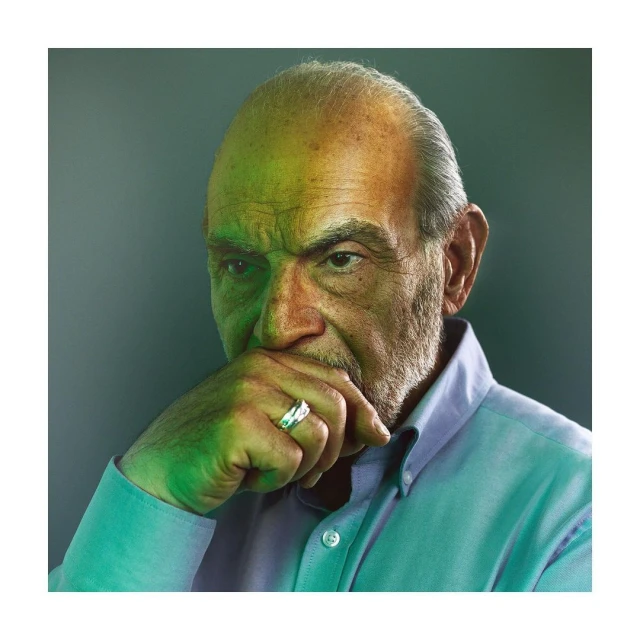 Portrait of an elderly man with a hand to his face, wearing a collared shirt, ring on finger; green and yellow cast lighting.