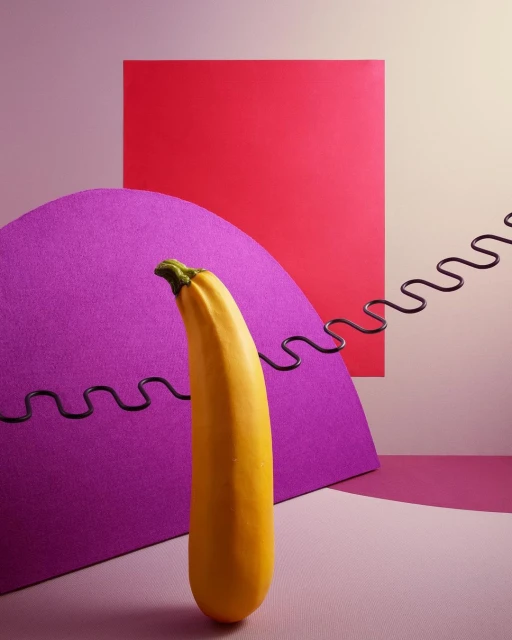 Still life with a yellow zucchini standing upright against a purple geometric background with a red square and black wire accent.