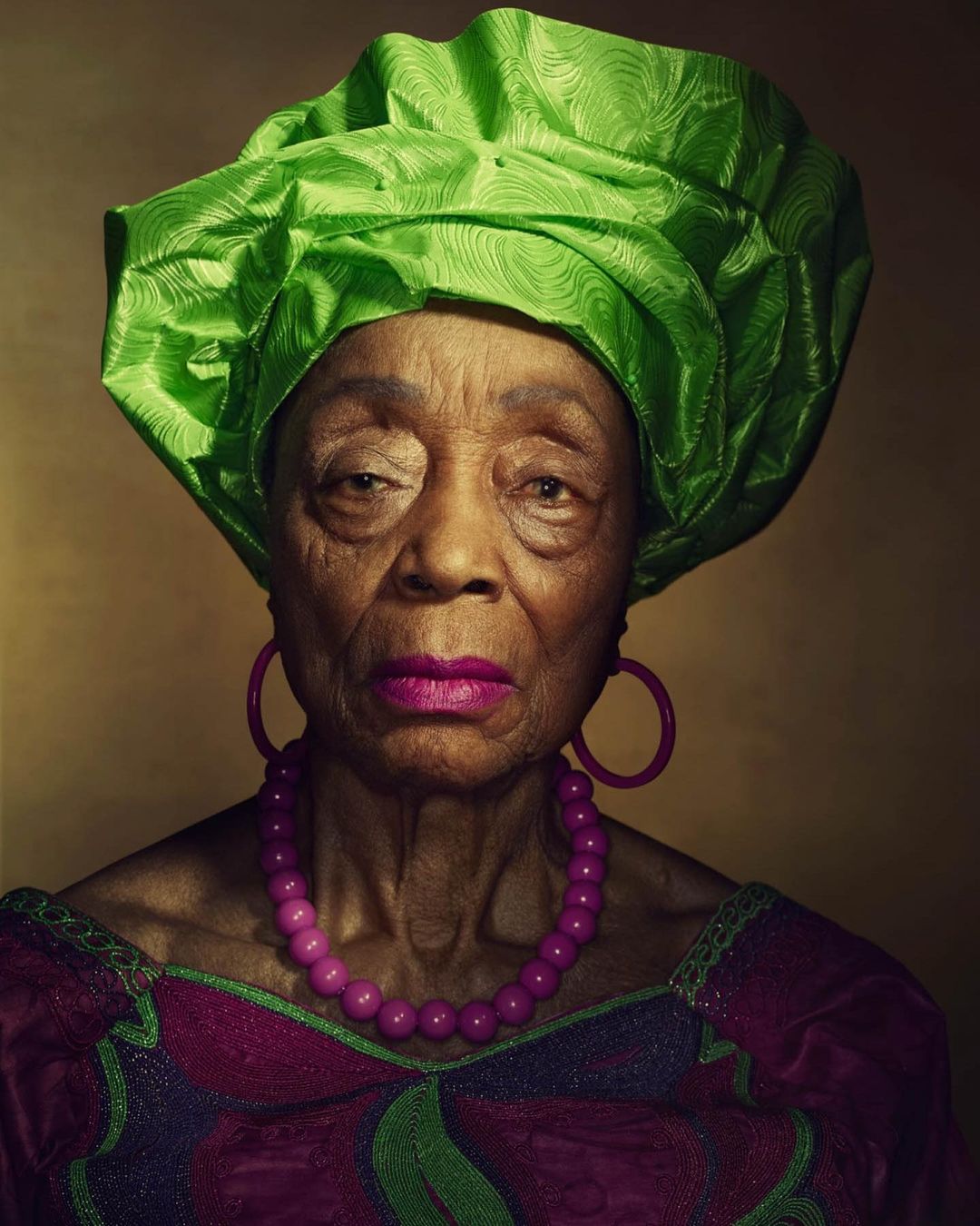 Close-up portrait of an elderly Black woman with a green headwrap, magenta lipstick, and pink jewelry, set against a warm brown background.