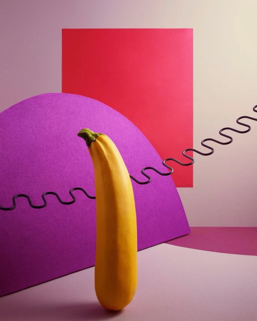 Vibrant still life featuring a yellow zucchini set against geometric shapes in shades of pink, purple, and a black spring.