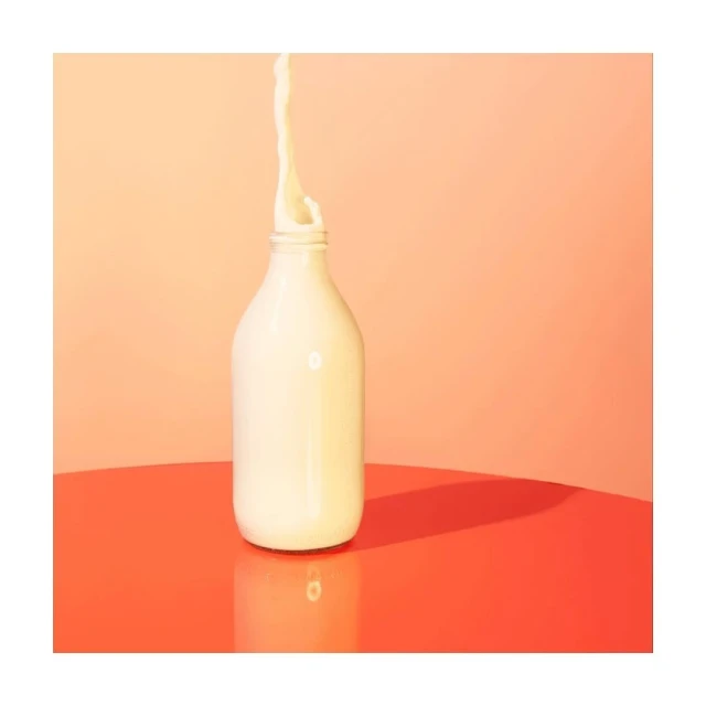 Glass milk bottle with milk splash on a coral table against a peach backdrop in a studio setting, showcasing a clean and refreshing presentation.