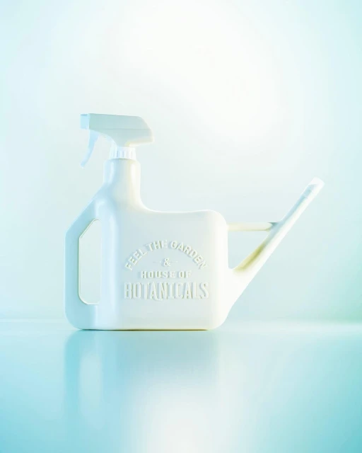 A white plastic watering can with a spray nozzle sits on a reflective surface against a pale background, casting a soft reflection.