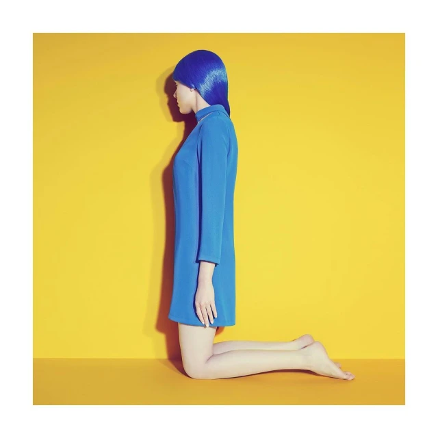 Side-view shot of a fair-skinned woman kneeling, wearing a vibrant blue dress and a matching blue wig, set against a solid yellow backdrop.