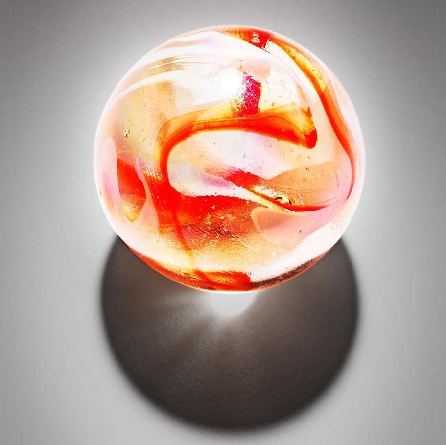 Close-up of a translucent orange and white swirl marble casting a shadow on a grey surface in a studio shot. Toy, collectible.
