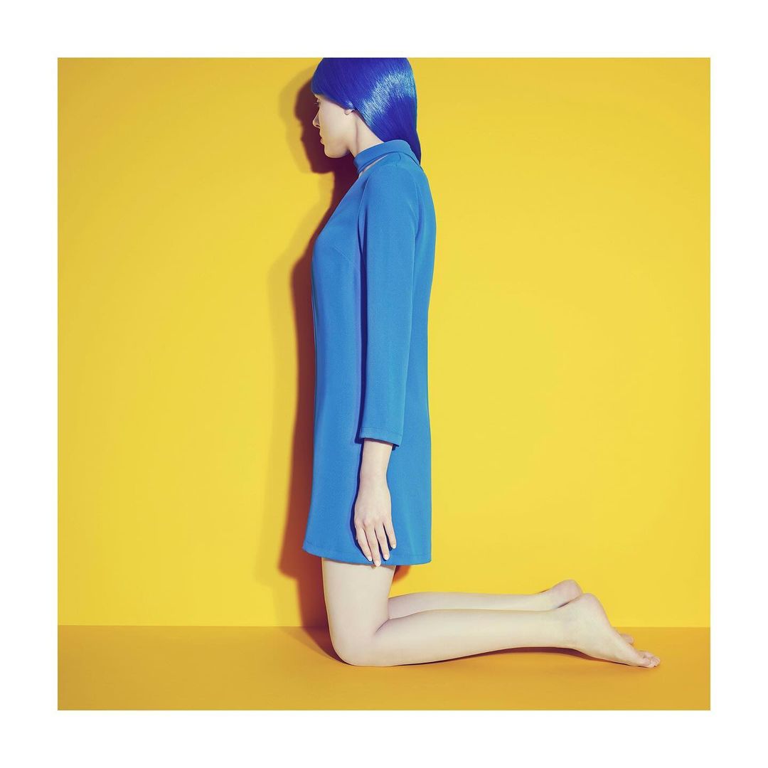 Side view of a person kneeling in a blue dress and matching wig against a yellow background, studio shot, fashion-forward, minimalist.
