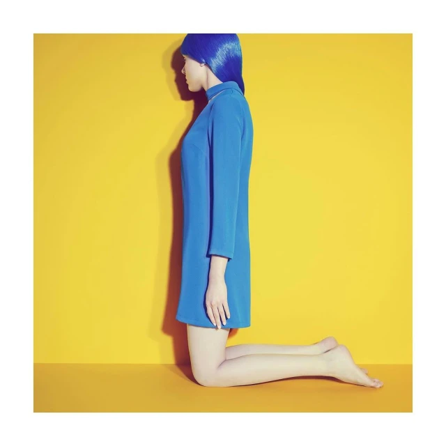 Side view of a person kneeling in a blue dress and matching wig against a yellow background, studio shot, fashion-forward, minimalist.