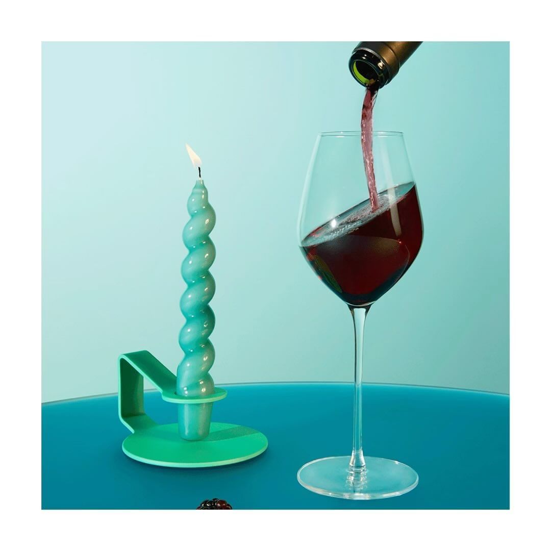 Wine pours into a glass beside a burning teal candle on a matching teal table against a light teal backdrop in an indoor setting.