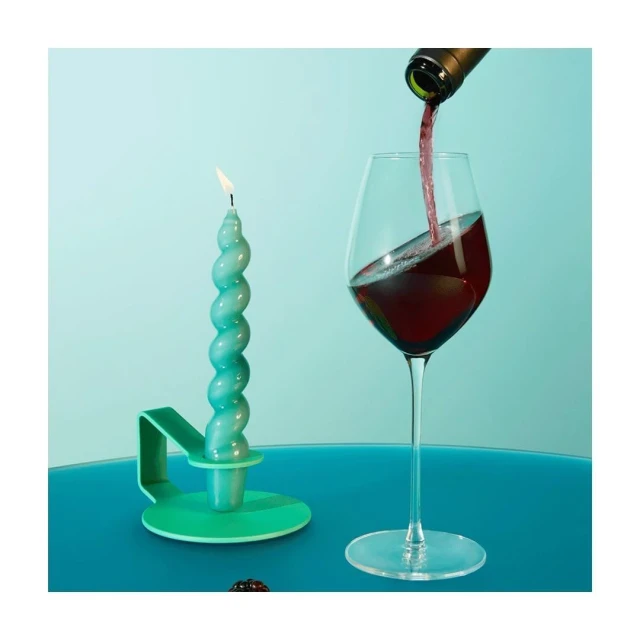Wine pours into a glass beside a burning teal candle on a matching teal table against a light teal backdrop in an indoor setting.