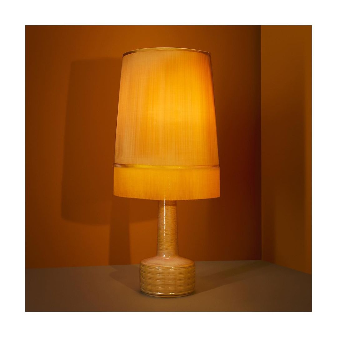 Illuminated table lamp in amber tones with a cylindrical shade and textured base, casting soft light against a golden-brown wall.