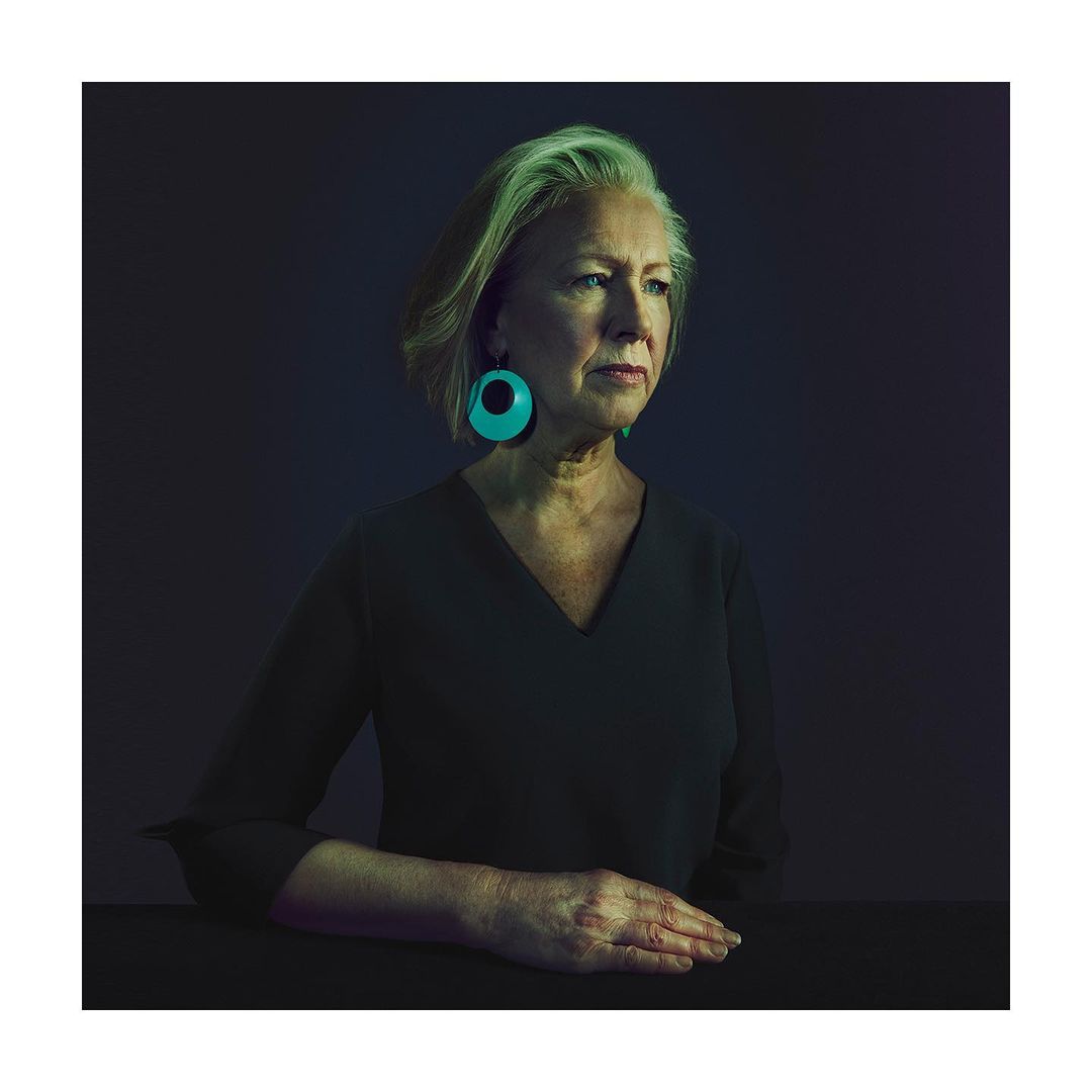 Portrait of a mature woman with short grey hair wearing green earrings and a black v-neck dress against a dark background.
