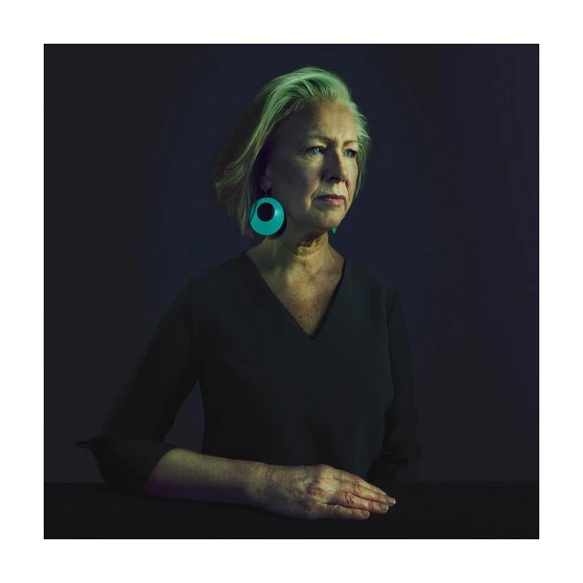 Portrait of a mature woman with short grey hair wearing green earrings and a black v-neck dress against a dark background.