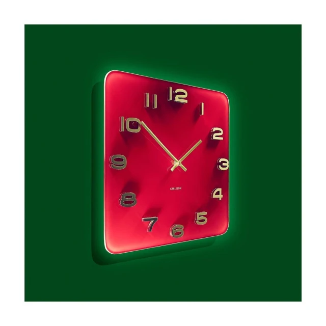 Red square clock displaying time with gold hands and numerals set against a dark green background in a studio setting.