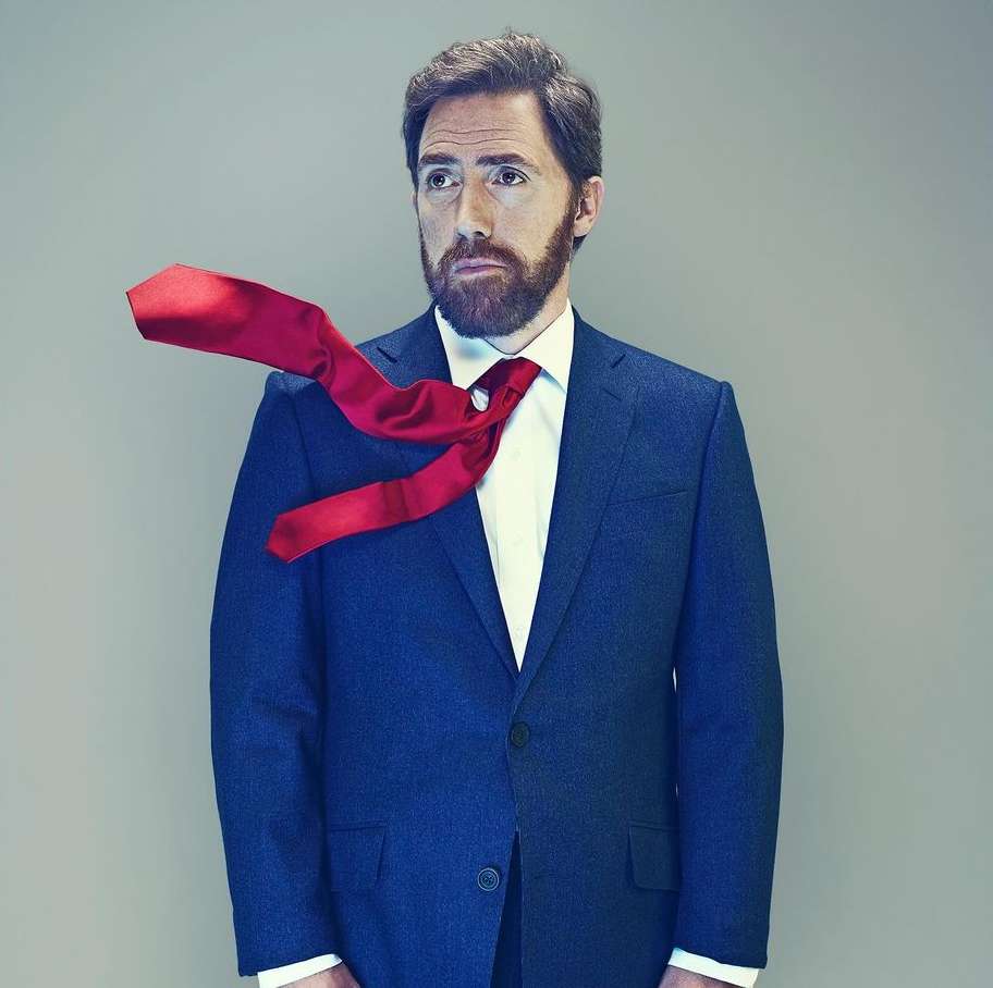 Portrait of an adult male with a beard wearing a blue suit and white shirt, with a red tie flying off to the side against a gray backdrop.