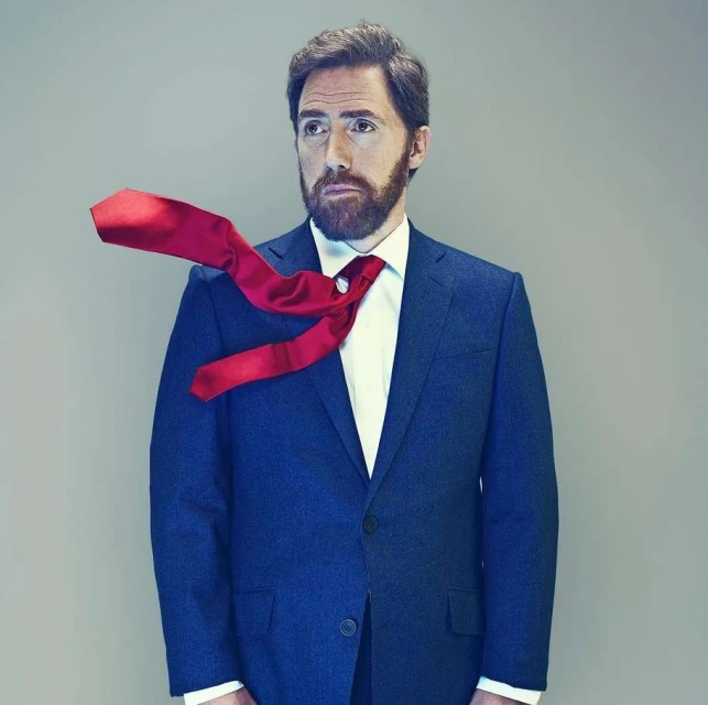 Portrait of an adult male with a beard wearing a blue suit and white shirt, with a red tie flying off to the side against a gray backdrop.
