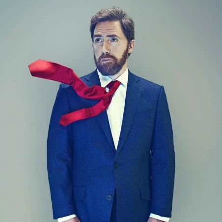 Portrait of an adult male with a beard wearing a blue suit and white shirt, with a red tie flying off to the side against a gray backdrop.