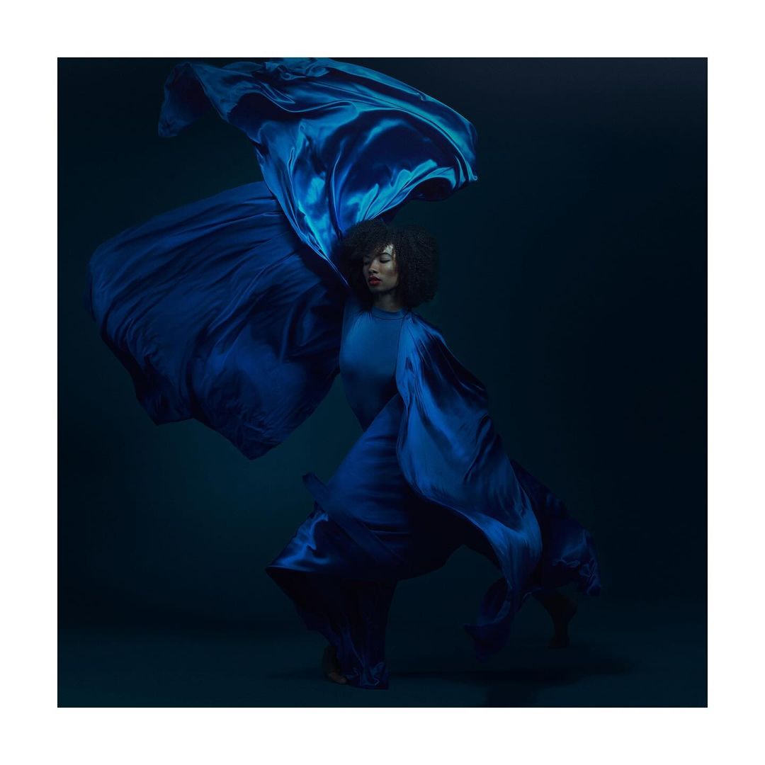 Adult woman with curly hair and flowing blue fabric poses in a portrait with a dark background in a fashion photography style.