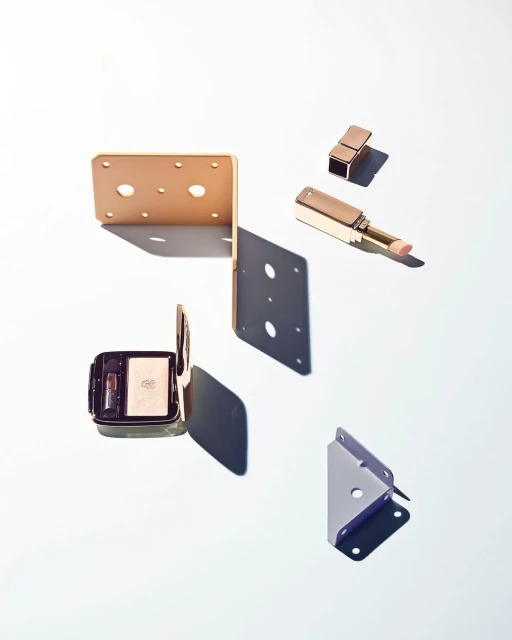Still life shot of gold lipstick, eye shadow makeup and geometric corner brackets on white, showcasing beauty and hardware objects.