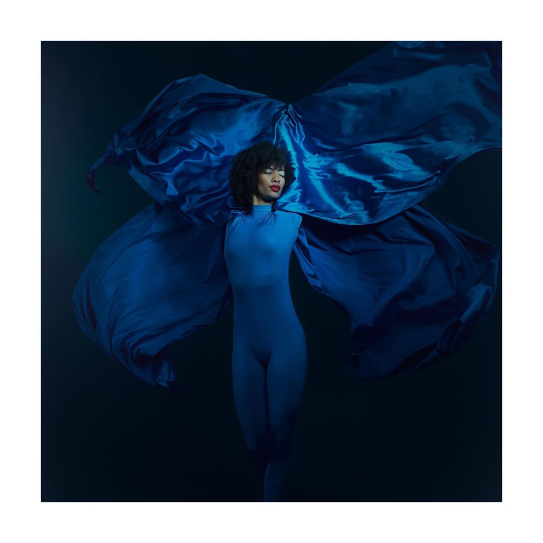 Elegant woman in blue bodysuit poses dramatically with flowing satin fabric in a studio portrait against a dark background with spotlight.