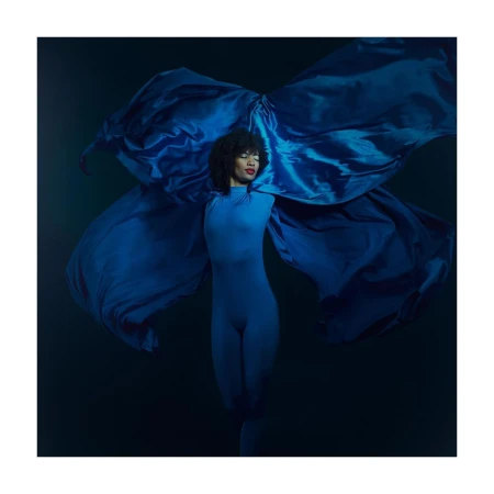 Elegant woman in blue bodysuit poses dramatically with flowing satin fabric in a studio portrait against a dark background with spotlight.