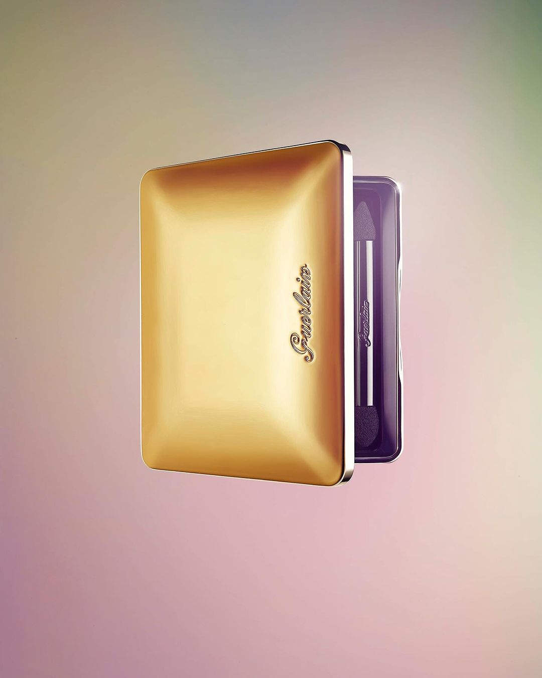 Golden Guerlain eyeshadow palette with applicator brush on a pink and green gradient background, showcasing makeup and cosmetic product.