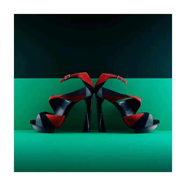 Two high-heeled platform shoes with red and black suede straps are mirrored against a split dark green and black background.