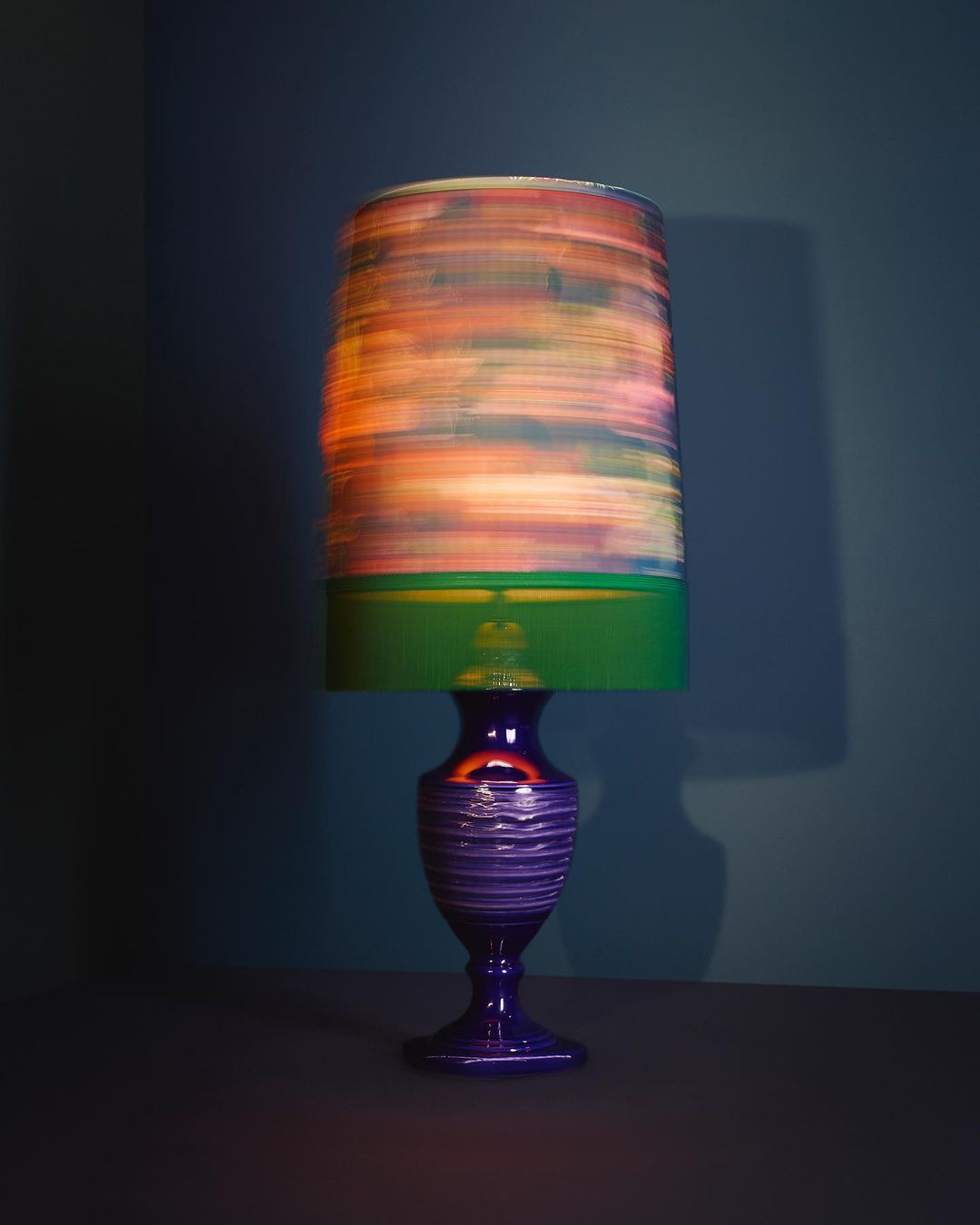 Lamp with colorful shade and blue glass base sitting on a dark table against a blue wall in a room corner, interior lighting.
