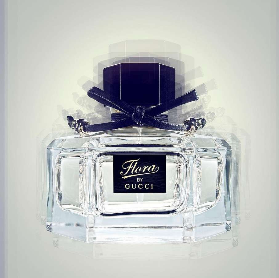 Close-up of a glass perfume bottle with a black cap and ribbon, showcasing the 'Flora by Gucci' label against a white background.