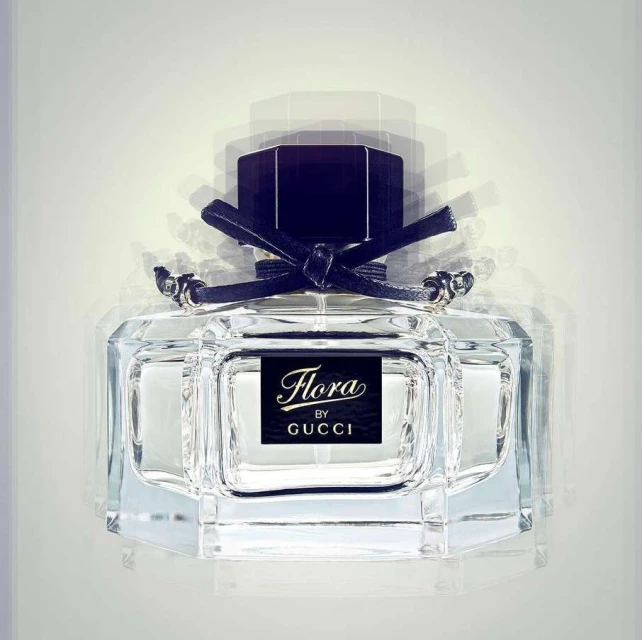 Close-up of a glass perfume bottle with a black cap and ribbon, showcasing the 'Flora by Gucci' label against a white background.