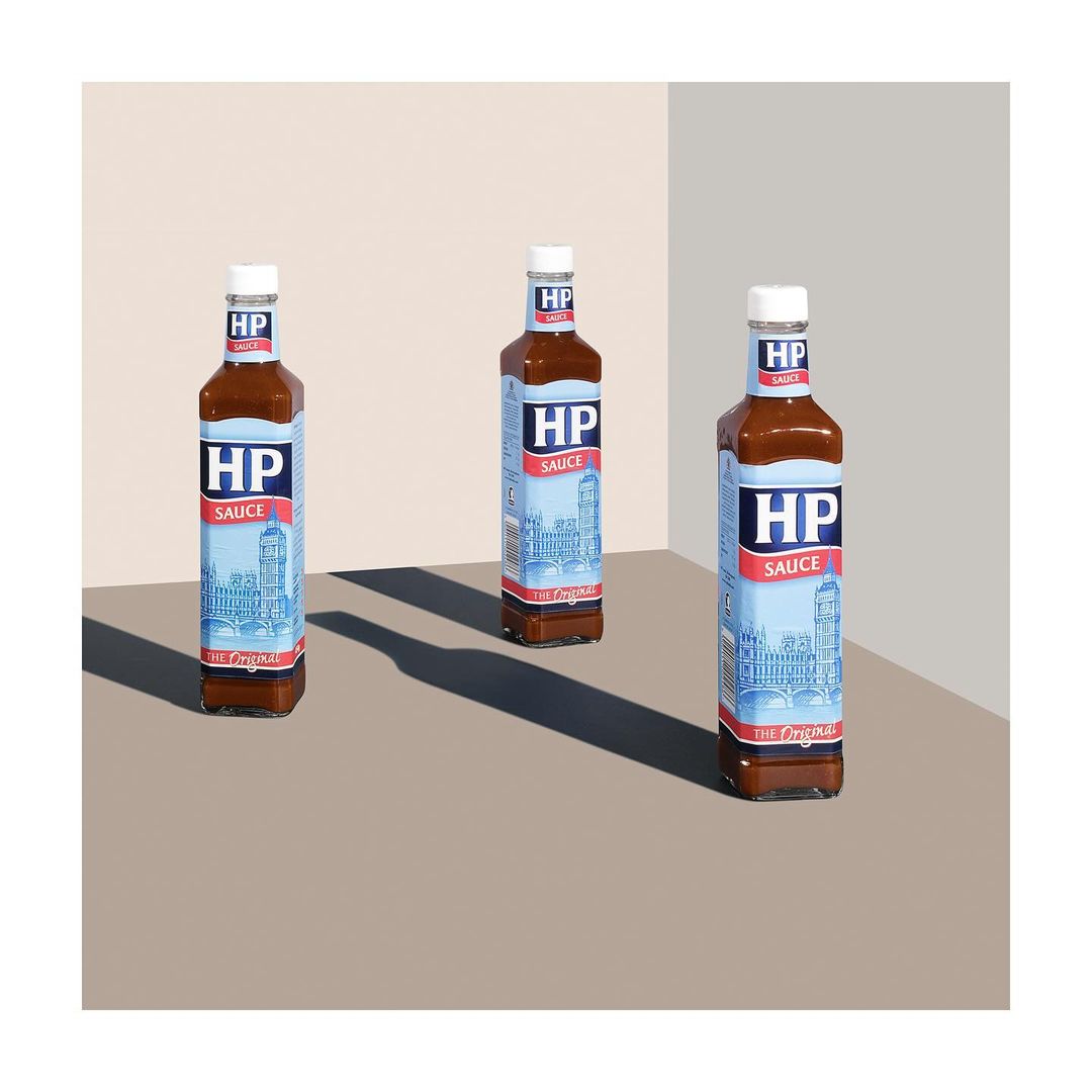 Three bottles of HP Sauce are arranged on a beige surface, casting dark shadows in a well-lit, minimalist studio setup. Food photography.