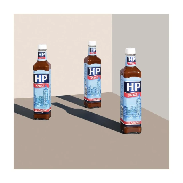 Three bottles of HP Sauce are arranged on a beige surface, casting dark shadows in a well-lit, minimalist studio setup. Food photography.