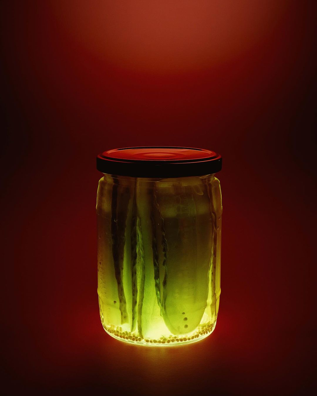 A jar of green pickled cucumbers is backlit against a dark red background, casting a yellow glow and showcasing the texture of the glass.