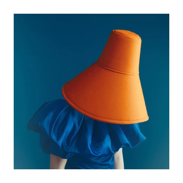 Person wearing a blue ruffled top and a large, orange, cone-shaped hat against a plain blue backdrop in a studio setting.