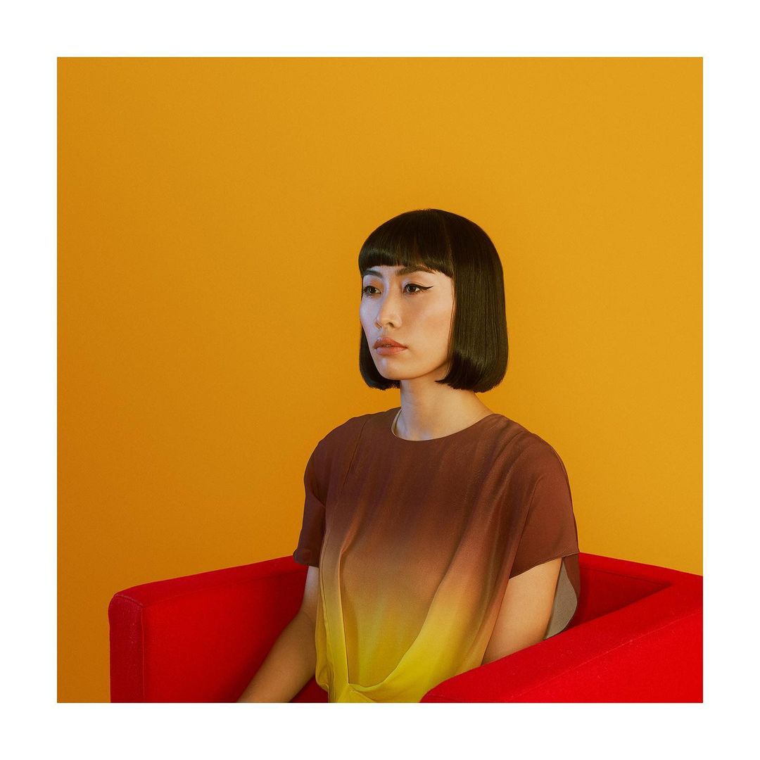Woman with a modern bob haircut and brown and yellow top posing against a yellow backdrop while sitting in a red chair.