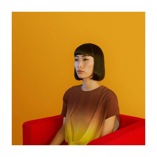 Woman with a modern bob haircut and brown and yellow top posing against a yellow backdrop while sitting in a red chair.