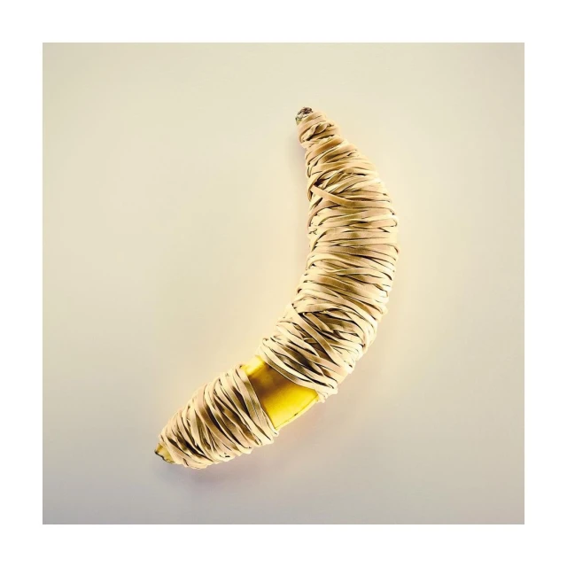 Banana covered in tan rubber bands against an off-white background in a studio shot. Food photography featuring textures and creative food art.