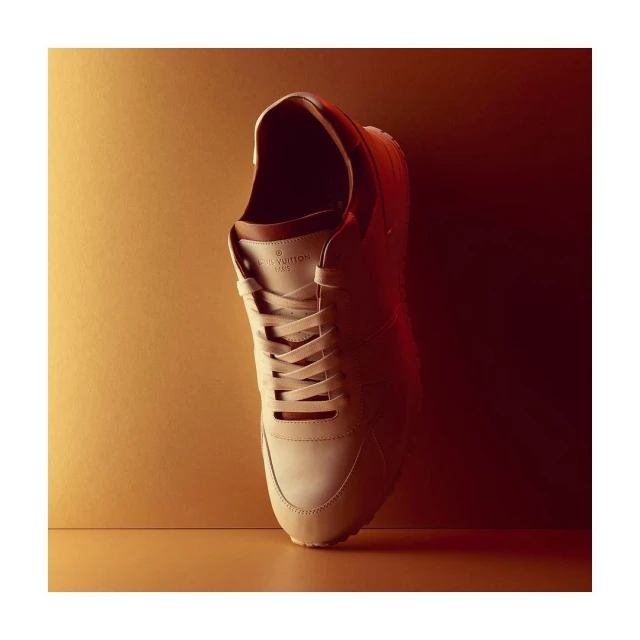 Studio shot of a tan-colored Louis Vuitton sneaker with brown trim, laces, and white soles, positioned against a background with gradient lighting.