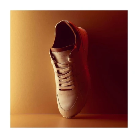 Studio shot of a tan-colored Louis Vuitton sneaker with brown trim, laces, and white soles, positioned against a background with gradient lighting.