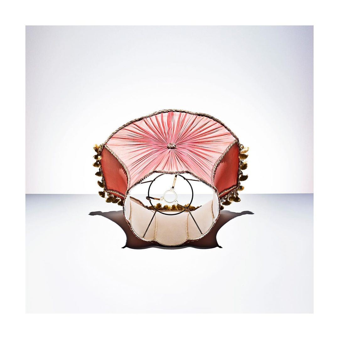 Close-up shot of an ornate lampshade with a salmon pink fabric shade and ivory interior, casting a soft shadow on a white background.