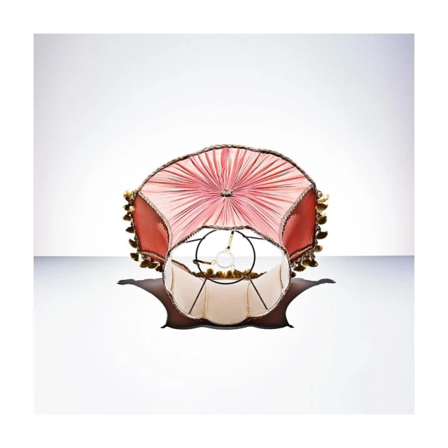 Close-up shot of an ornate lampshade with a salmon pink fabric shade and ivory interior, casting a soft shadow on a white background.
