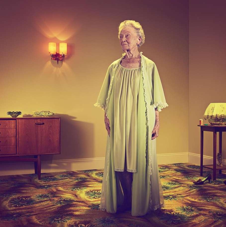 Elderly woman in a light green robe stands in a vintage-style room with floral carpet, cabinet, table lamp, and decorative objects.