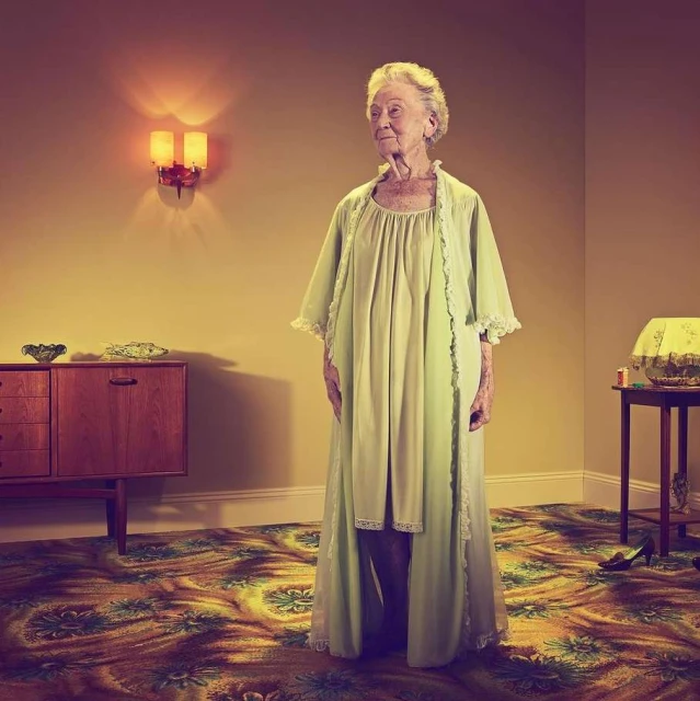 Elderly woman in a light green robe stands in a vintage-style room with floral carpet, cabinet, table lamp, and decorative objects.