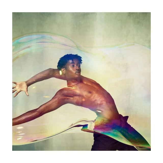 African American male with arm outstretched surrounded by large iridescent soap bubbles in a light-filled, textured studio setting.