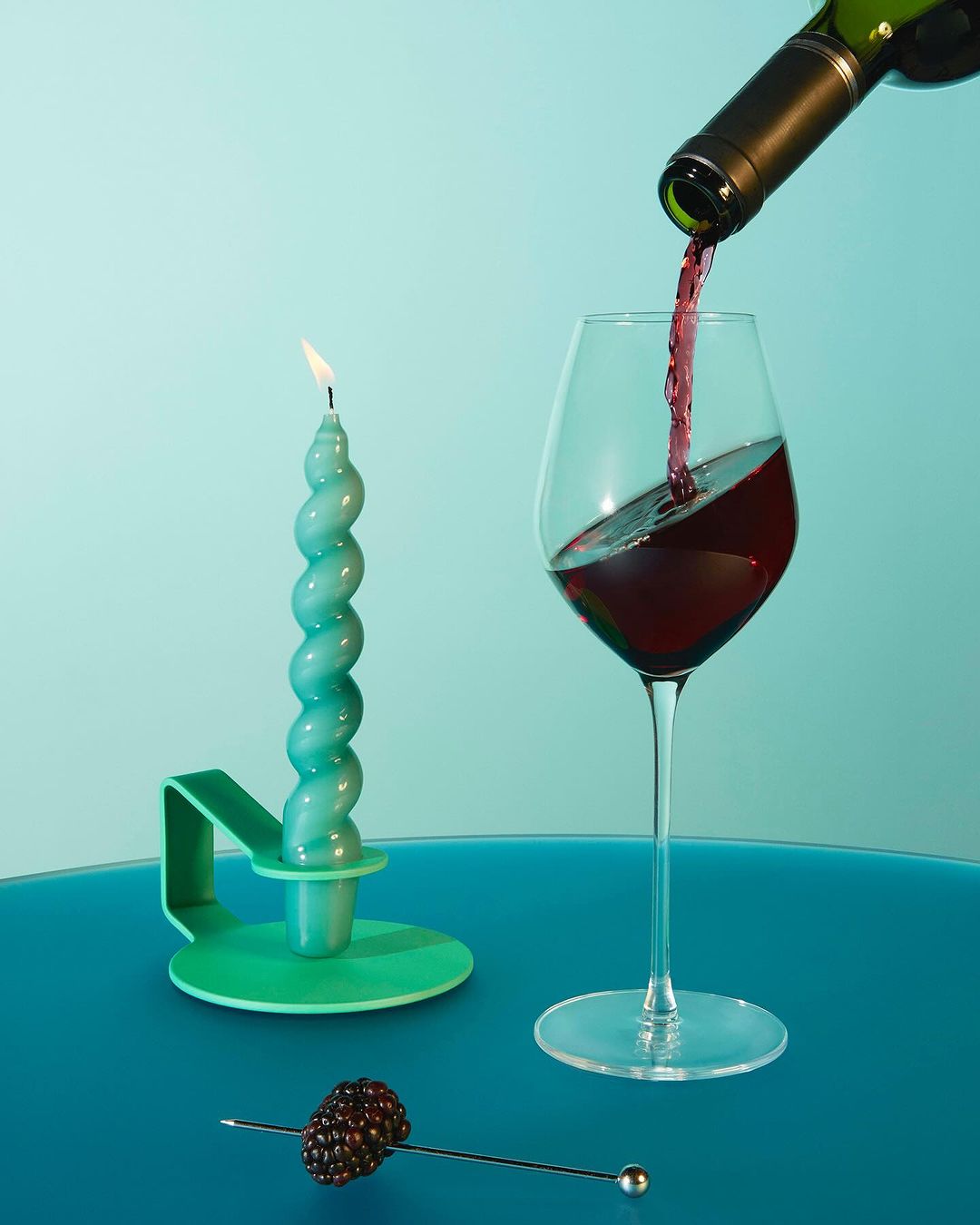 Wine pouring into a glass next to a candle and a blackberry garnish, set against a light blue background. Still life shot.
