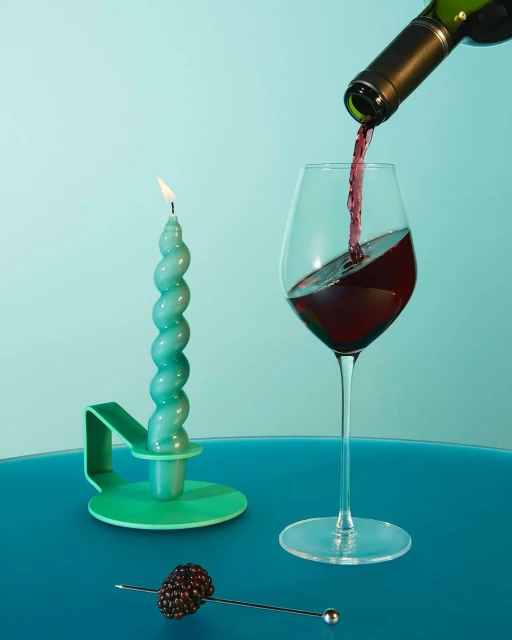 Wine pouring into a glass next to a candle and a blackberry garnish, set against a light blue background. Still life shot.