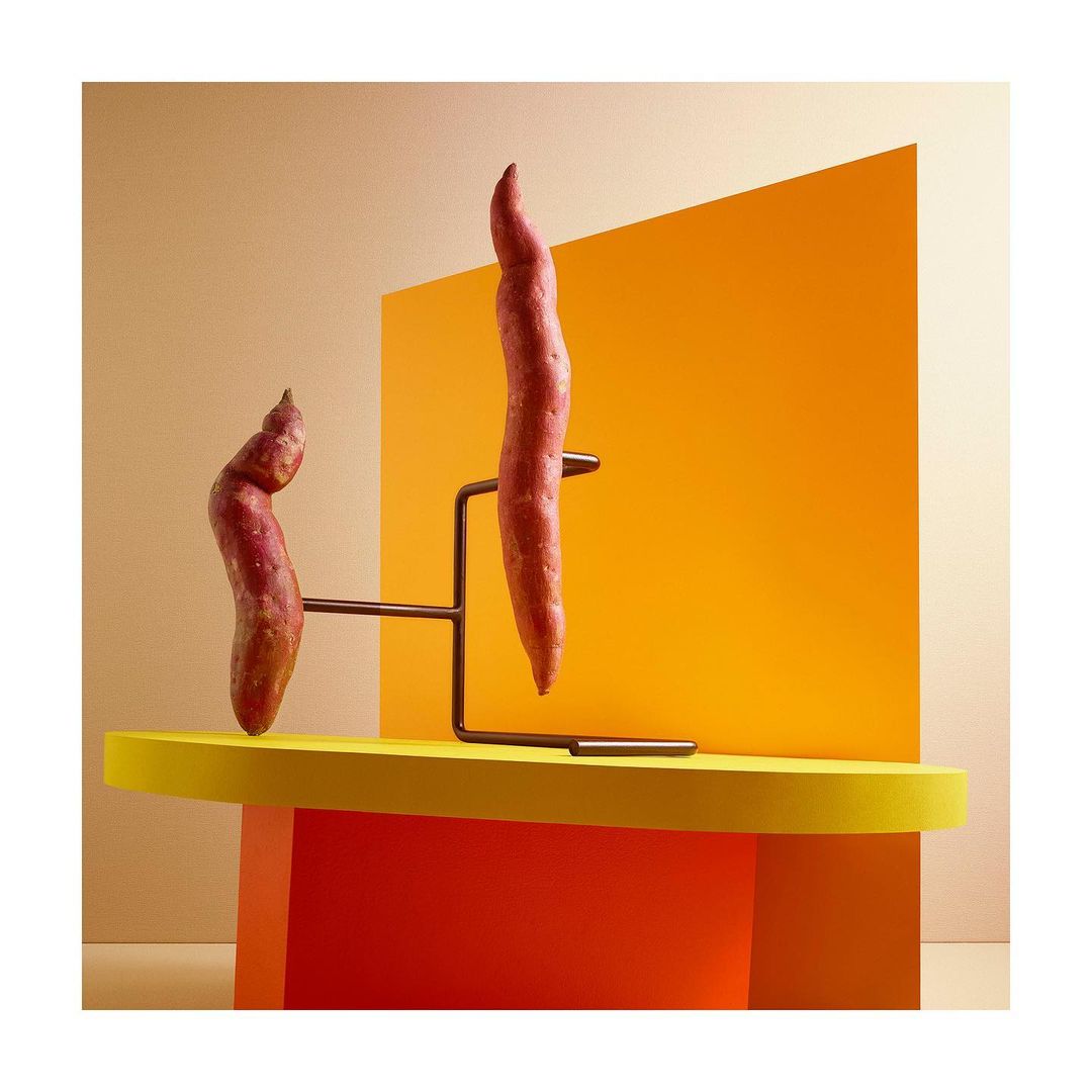 Still life studio shot of two sweet potatoes presented on a modern metal stand against a split beige and orange color block background.