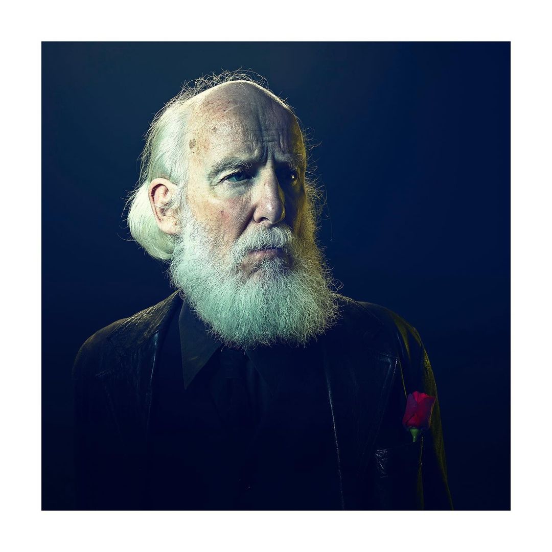 Portrait of a mature adult man with a long white beard wearing a dark jacket and a rose, set against a dark blue background.