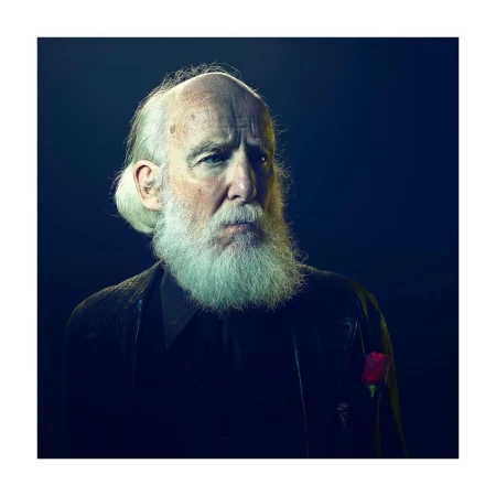 Portrait of a mature adult man with a long white beard wearing a dark jacket and a rose, set against a dark blue background.